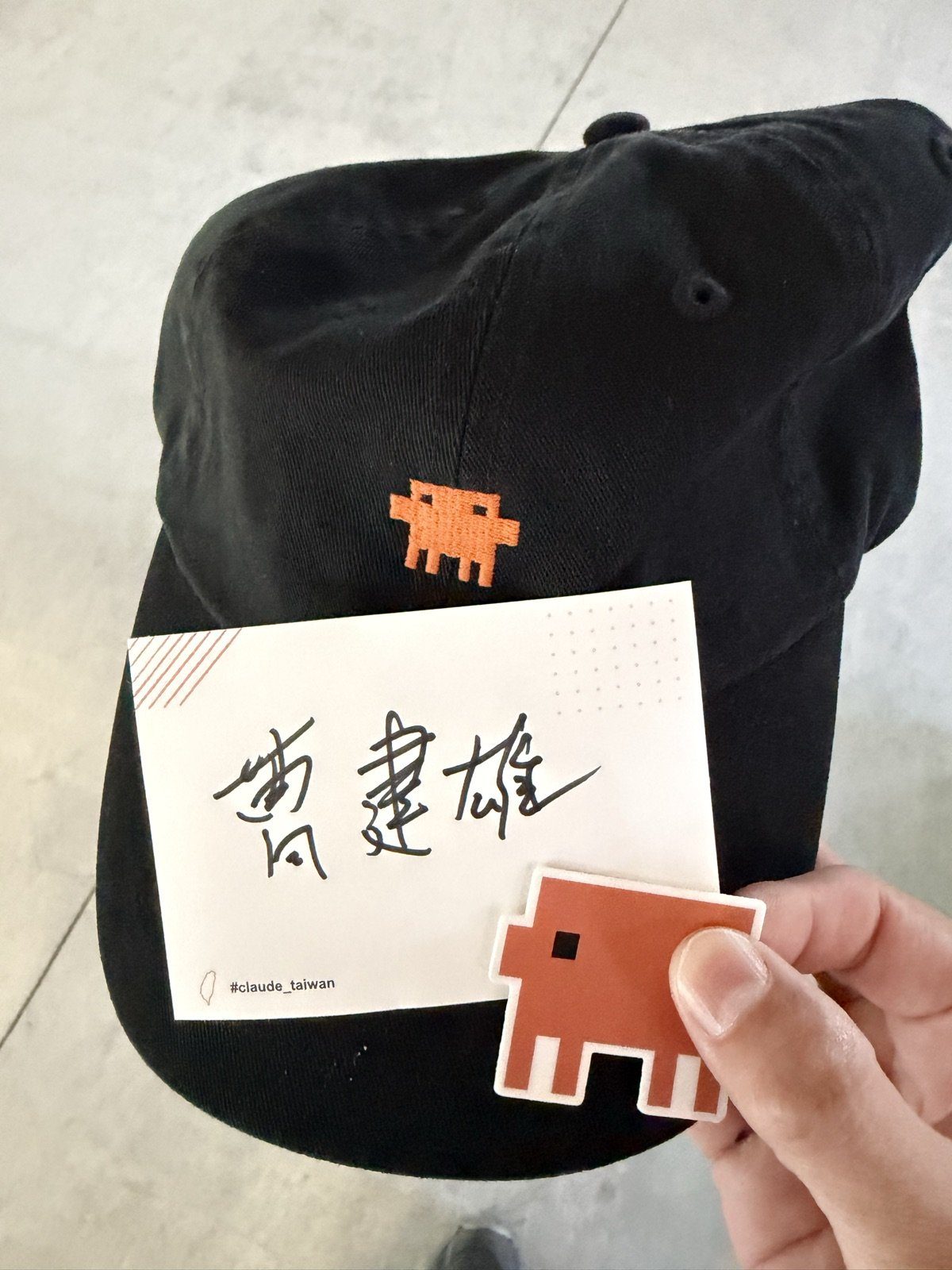 黑色Claude Code帽子，上面別著手寫「曹建雄」的名牌，手上拿著橘色像素小怪物貼紙